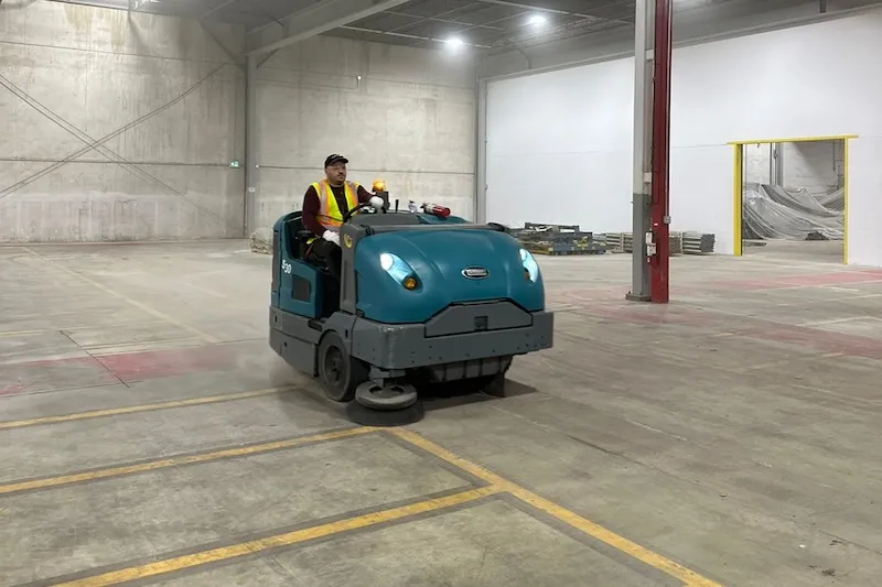 Operator using a ride-on sweeper to clean a large parking lot Technician operating a commercial ride-on sweeper machine to remove dust and debris from a concrete parking surface