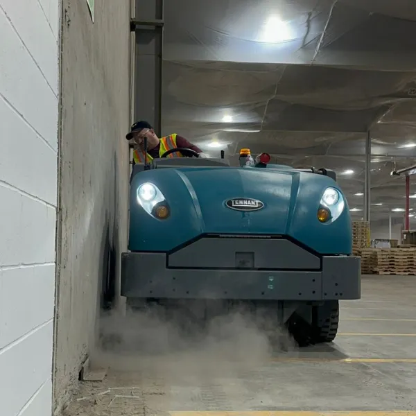 A worker operating a Tennant power sweeper to clean a warehouse floor near a wall.